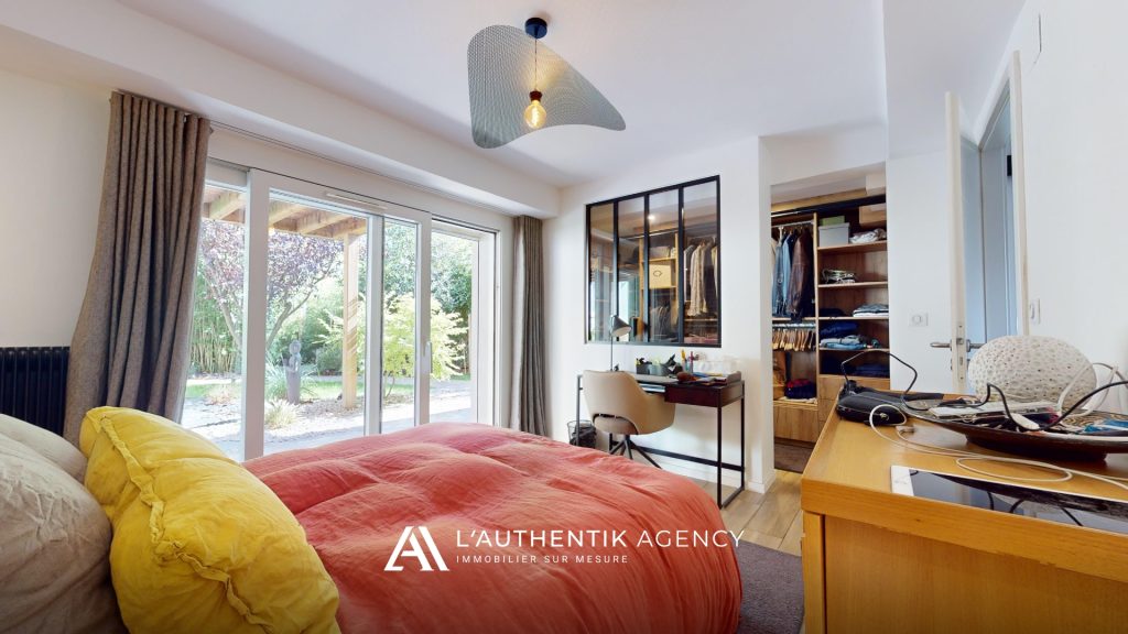 Vue depuis le chevet sur une chambre spacieuse avec baie vitrée donnant sur l’extérieur, un demi-mur avec verrière dévoile un dressing de belle taille, et un bureau complète harmonieusement l’aménagement.