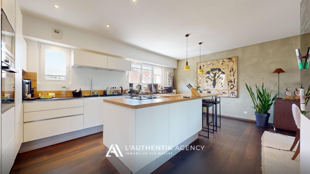 Cuisine blanche ouverte et lumineuse avec plans de travail en bois clair. Elle est dotée de nombreux rangements et d’un îlot central, alliant esthétique épurée et fonctionnalité.